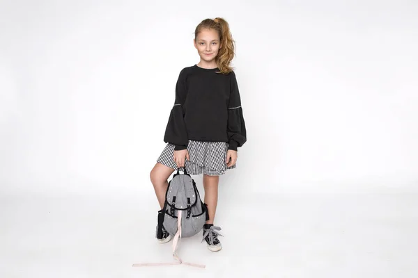 Linda niña vestida con ropa de escuela y mochila escolar sentada sobre fondo blanco —  Fotos de Stock