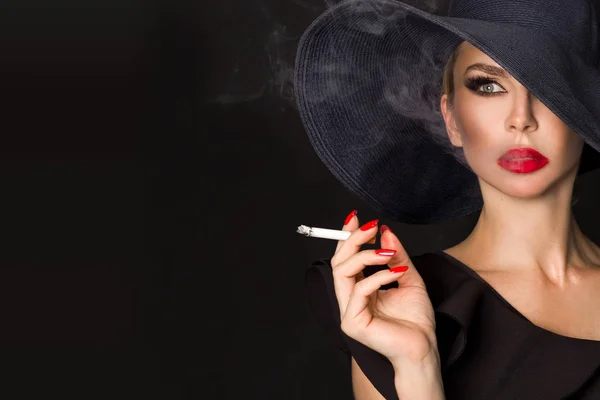 エレガントな女性は、黒のファム ・ ファタールはタバコを持つ手に帽子します。黒い背景に — ストック写真