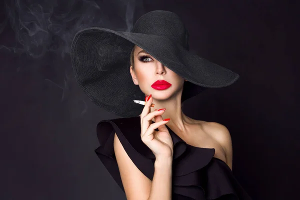 エレガントな女性は、黒のファム ・ ファタールはタバコを持つ手に帽子します。黒い背景に — ストック写真