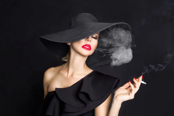 Mujer elegante, femme fatale en sombrero negro con cigarrillo en la mano. Sobre fondo negro — Foto de Stock