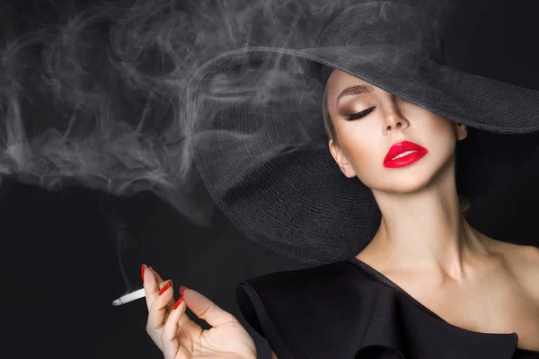 Mujer elegante, femme fatale en sombrero negro con cigarrillo en la mano. Sobre fondo negro — Foto de Stock