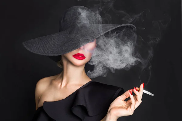 Mujer elegante, femme fatale en sombrero negro con cigarrillo en la mano. Sobre fondo negro — Foto de Stock