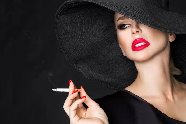 Donna elegante, femme fatale in cappello nero con sigaretta in mano. Su sfondo nero — Foto Stock