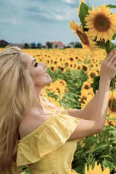 Mulher loira bonita de pé em um prado com girassóis e sorrindo feliz — Fotografia de Stock