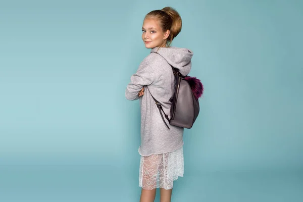 Симпатична маленька дівчинка одягнена в шкільний одяг і шкільний рюкзак, що стоїть на синьому фоні — стокове фото