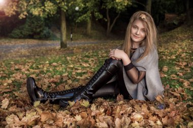 Sevimli küçük kız ceket giymiş ve çizmeler, sonbahar arka plan üzerinde oturan altın sonbahar yaprakları.