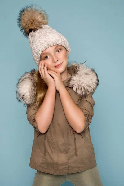 Söt liten flicka klädd i vinterkläder, hatt och vinter jacka med päls, stående på blå bakgrund — Stockfoto