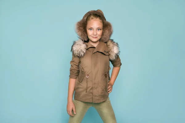 Симпатична дівчинка, одягнена в зимовий одяг, і зимова куртка з хутром, стоїть на синьому фоні — стокове фото