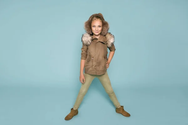 Linda niña vestida con ropa de invierno, sombrero y chaqueta de invierno con piel, de pie sobre fondo azul — Foto de Stock