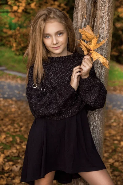 Roztomilá holčička na sobě kabát a boty sedí na podzimní pozadí, zlaté podzimní listy. — Stock fotografie