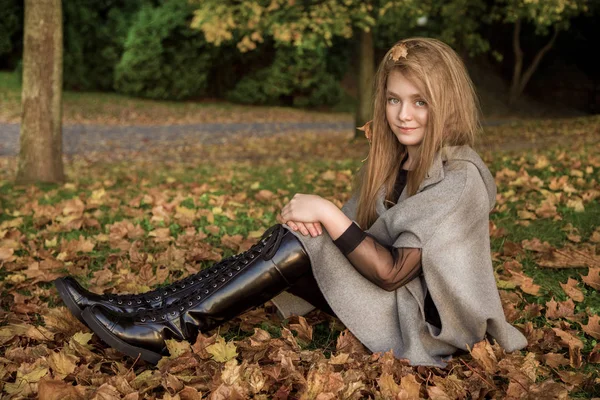 Linda niña vestida con abrigo y botas sentadas sobre fondo de otoño, hojas doradas de otoño . — Foto de Stock