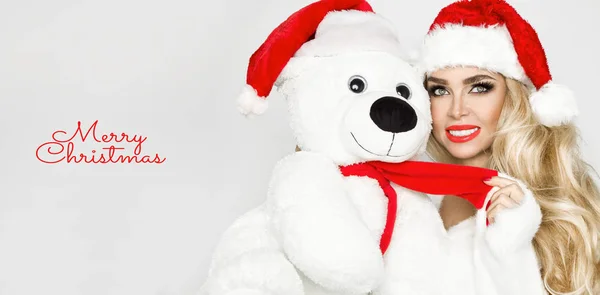 Belle mannequin féminine blonde sexy vêtue d'un chapeau du Père Noël embrasse un ours en peluche blanc dans une casquette rouge. Sensuelle fille pour Noël . — Photo