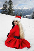 Krásná sexy blondýnka na sobě červené šaty a klobouk Santa na pozadí hor a sněhu. Krásná, smyslná žena, Santa Claus