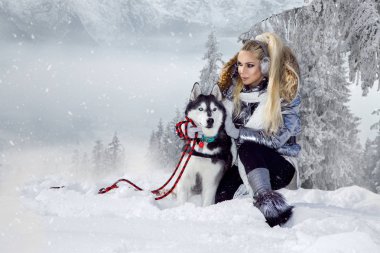Güzel sarışın kadın, kar üzerinde duran ve Husky köpek tutarak. Arka planda dağlar ve kar güzel bir görünümü vardır.