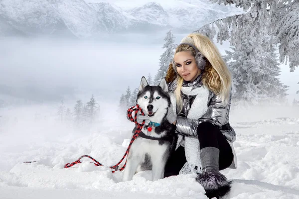 Güzel sarışın kadın, kar üzerinde duran ve Husky köpek tutarak. Arka planda dağlar ve kar güzel bir görünümü vardır.