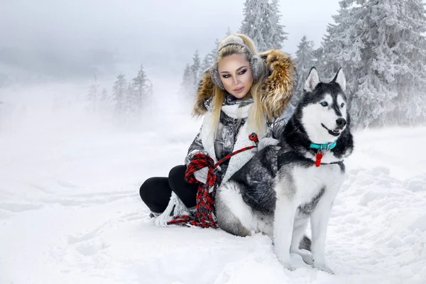 Güzel sarışın kadın, kar üzerinde duran ve Husky köpek tutarak. Arka planda dağlar ve kar güzel bir görünümü vardır.