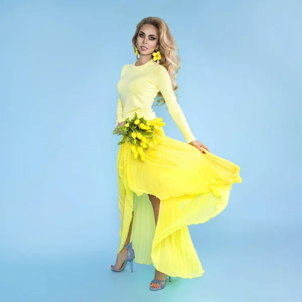 Spring fashion Woman. Beautiful model with colorful clothes, holding a bouquet of spring flowers posing over blue background . Fashion photo. Easter concept.
