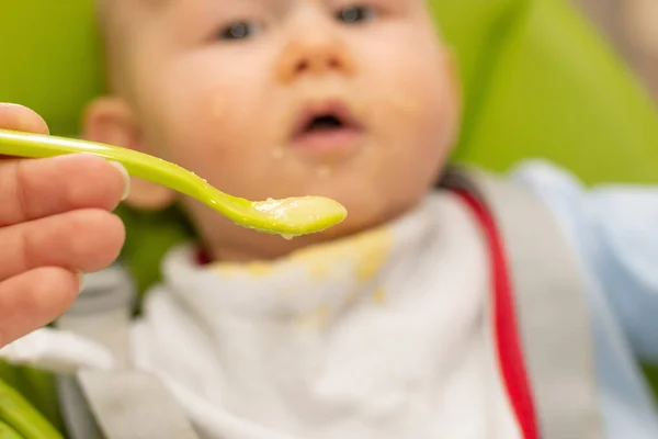 Colher cheia de mingau de cereal e bebê embaçado no fundo. Foco seletivo na colher. Bebê primeiro soilds. Desmame . — Fotografia de Stock