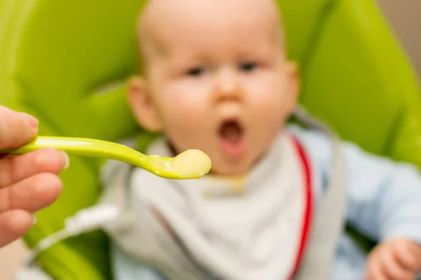 Colher cheia de mingau e bebê embaçado com a boca aberta no fundo. Foco seletivo na colher. Bebê primeiro soilds. Desmame . — Fotografia de Stock