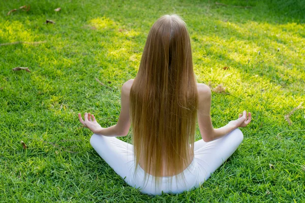 Молодая красивая девушка-подросток с длинными светлыми волосами медитирует в парке на зеленой траве — стоковое фото