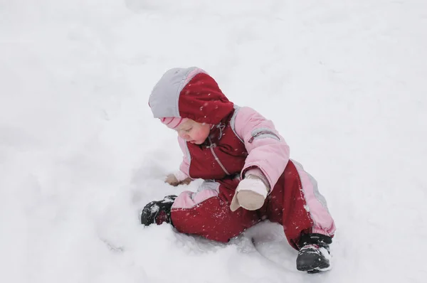 Inverno no parque. Uma menina de criança vestindo roupas quentes sentado no chão, que é coberto com neve — Fotografia de Stock