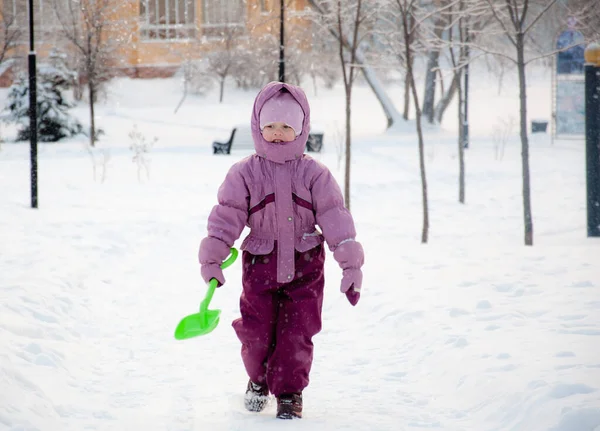 Inverno no parque. Uma menina usando roupas quentes andando pelo snowfalll no parque segurando uma pá verde em sua mão — Fotografia de Stock