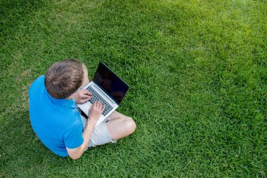 Parktaki yeşil çimlerde dizüstü bilgisayarıyla oturan bir adam. Dışarıda çalışıyorum. Yukarıdan bak. Metin girdisi için boş boşluk