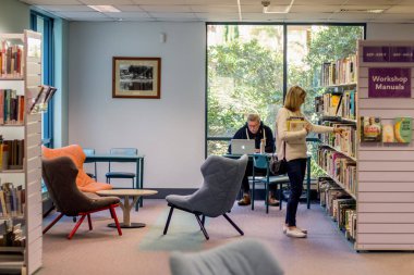Sydney, Avustralya - 2019-07-19 Yerel kütüphanede. Kitaplarla dolu kitap rafları ve okuyucular için sandalyeler. Dizüstü bilgisayar kullanan yaşlı bir adam ve ne okuyacağını seçen bir kadın. Sutherland, Nsw
