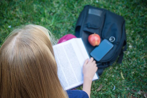 Student life concept. A girl with long blond hair sitting on a grass with the book on her knee. Her backpack and an apple for healthy snack laying nearby — Stock Photo, Image