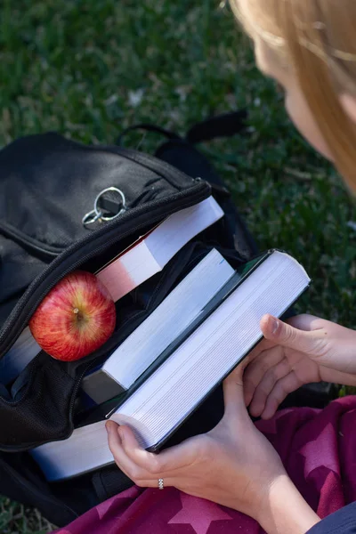 Student life concept. Black backpack stuffed with books. Hands holding books. Fresh apple for a healthy snack — Stock Photo, Image