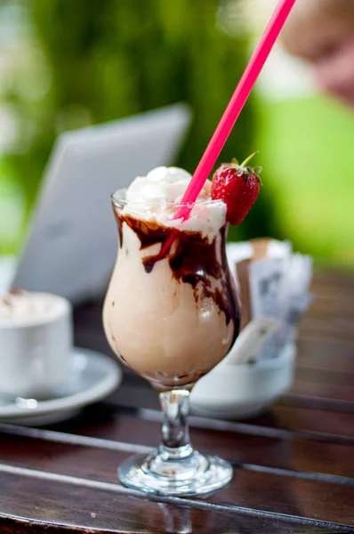 Um copo de frappe de batido de coquetel decorado com morango e chocklate na mesa de madeira com laptot e uma xícara de café no fundo. Conceito de férias — Fotografia de Stock