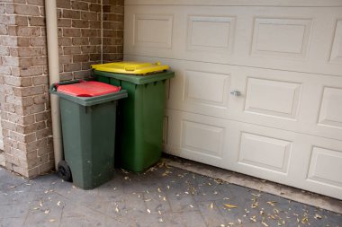 Australian garbage wheelie bins with red and yellow lids for general and recycling household waste near the resedential building garage door clipart