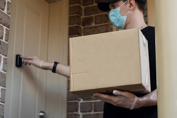 Contactless delivery during COVID-19 pandemic lockdown concept. Courier wearing mask and gloves holds a parcel with medical equipment and rings to the doorbell. Selective focus on a box