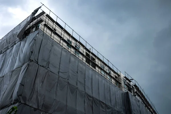 Building under construction with scaffolding and protective net is covered the building for safety and prevention dust and accidents against gray clouds before rainstorm.