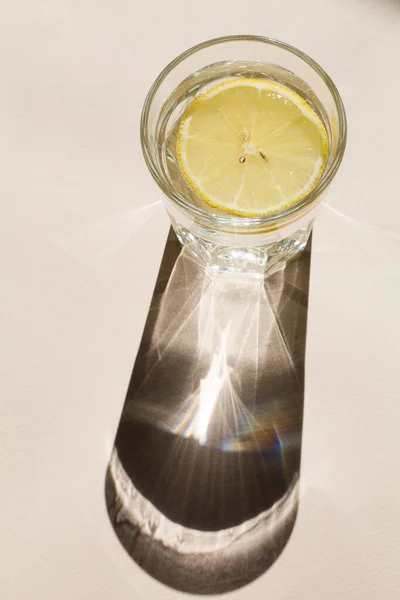 Top view glass of water with lemon slices on beige background with sunlight and deep shadow , summer lemonade, Ice drink, hot temperature