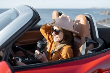 Elinde kahve fincanı olan bir kadın okyanus kıyısında bir araba kullanıyor.