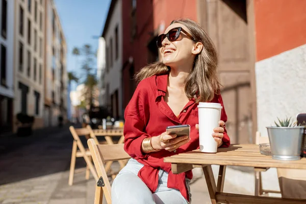 屋外のカフェテラスで電話とコーヒーを持つ女性 — ストック写真