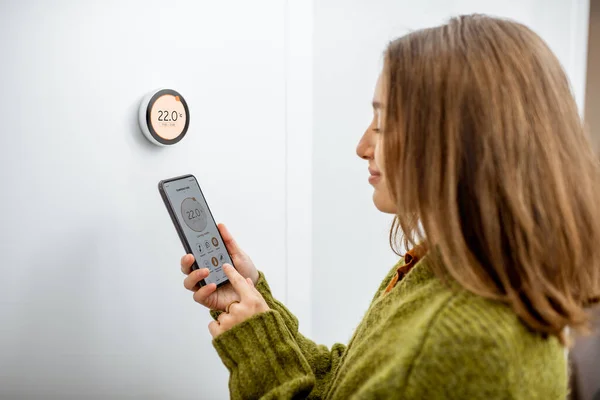 Donna che regola la temperatura di riscaldamento con telefono e termostato a casa — Foto Stock