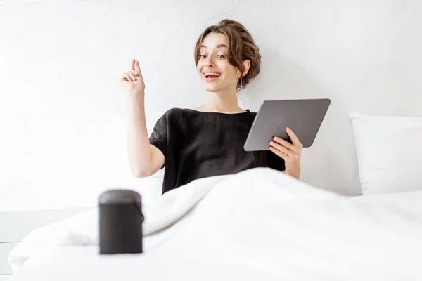 Mujer relajante con un altavoz inteligente y tableta en el dormitorio —  Fotos de Stock