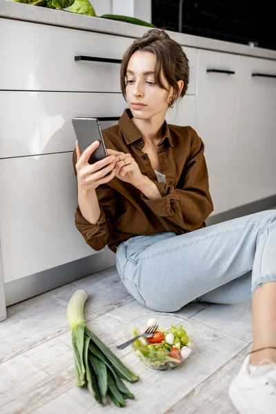 Mutfak zemininde sağlıklı yemek ve cep telefonu olan bir kadın. — Stok fotoğraf