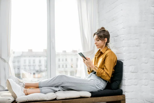 Žena pomocí telefonu, zatímco relaxovat na parapetu doma — Stock fotografie