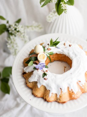 Kulich Geleneksel Paskalya tatlı ekmeği yeşil yapraklar, çiçekler ve beyaz yumurta ile süslenmiş beyaz kumaş arka planda beyaz bir tezgahta bir şeker.