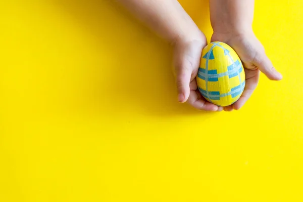 Рука закінчується. Великоднє жовте дерев'яне яйце з візерунком в руках дитини на жовтому тлі. вид зверху Стокова Картинка