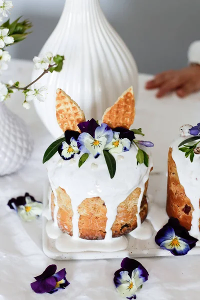 Paskalya pastası kulich. Geleneksel paskalya tatlı ekmeği tavşan kulakları ve leylak çiçekleriyle süslenmiş üç renkli Viola tabakta çiçek açan bir vazoya karşı. Boşluğu kopyala — Stok fotoğraf