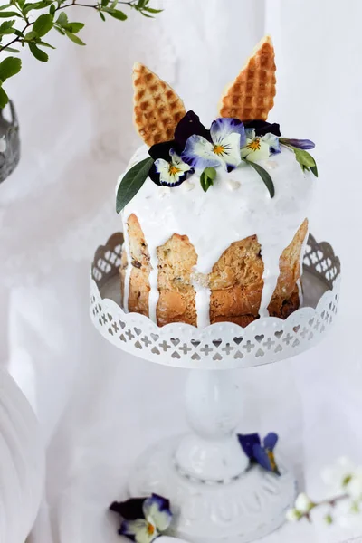 Torta pasquale kulich. Pane dolce tradizionale pasquale decorato con le orecchie di lepre e fiori lilla Viola tricolore su un piatto contro un vaso bianco con rami fioriti. Copia spazio — Foto Stock