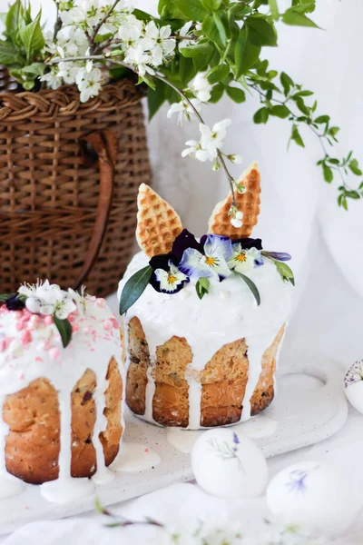 Paștele dulce kulich decorat cu urechi de iepure și flori de liliac Viola tricolor pe o placă albă de marmură pe fundalul unui coș de răchită cu ramuri cu flori și ouă de Paști . — Fotografie, imagine de stoc