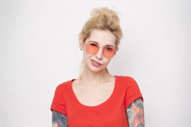 Genç belirsiz ve unassured sarışın kız portresi kırmızı rahat t-shirt giyiyor ve gözlük ret. Beyaz beton duvardan izole.