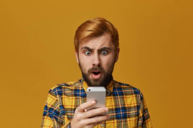 Portre yakışıklı redhead tıraşsız adam, boyalı akıllı telefon şok görünüyor. Teknoloji ve duygusal kavramı. Sarı arka plan üzerinde izole