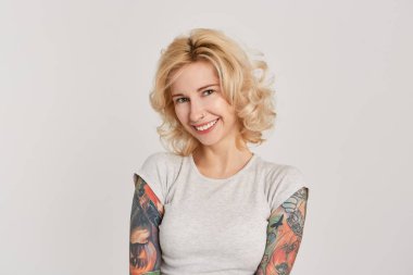 Burun ve tattoes kollarını doğrudan kamera seyir üzerinde Küpeli sarışın kız utangaç ve gülümseyerek. Beyaz duvardan izole