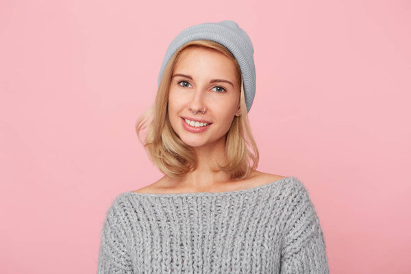 Portrait of young blonde woman, wears sweater and cap looks directly into camera and smiles. Shows her white teeth. Isolated over pink background